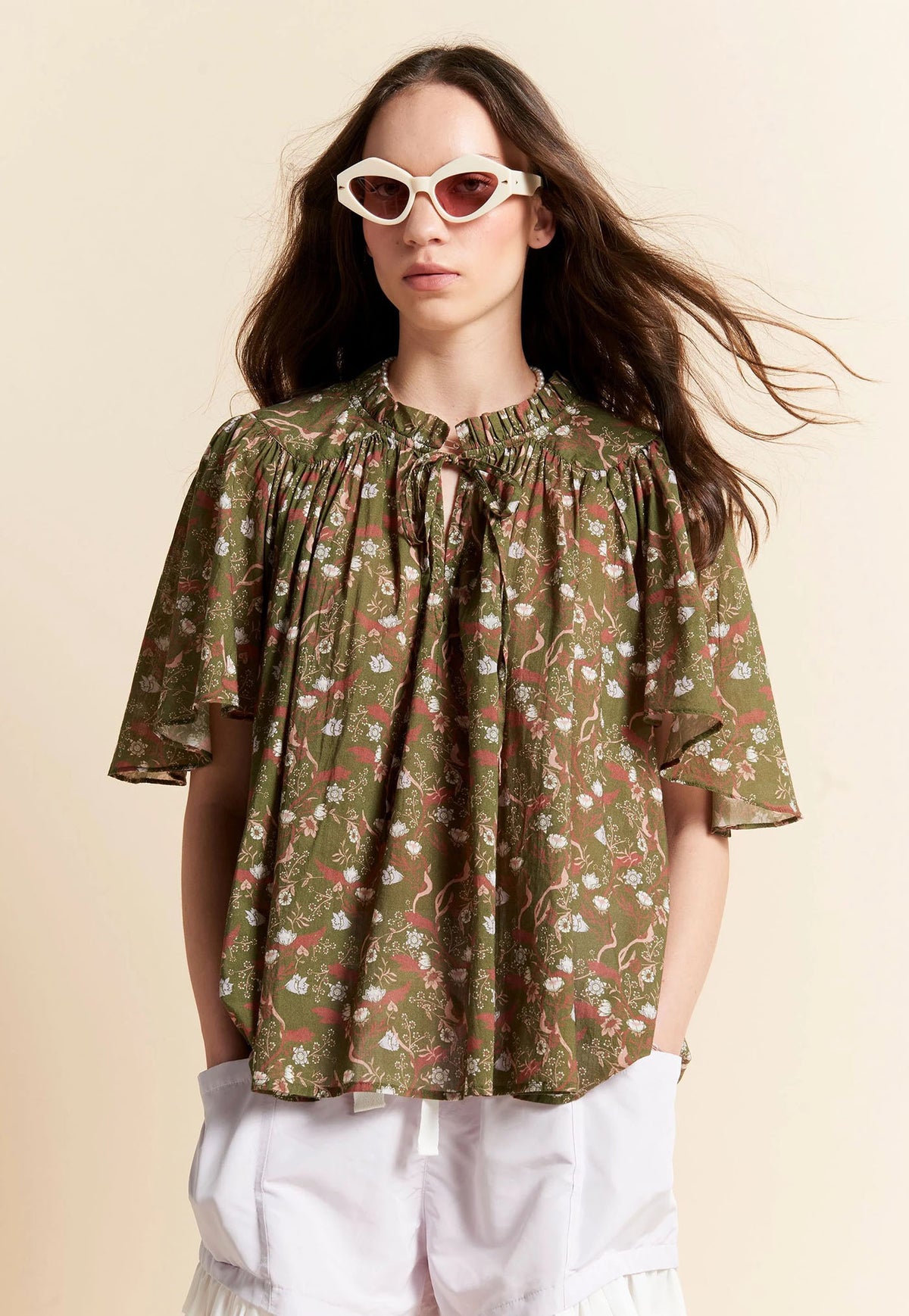 A woman wearing a short-sleeve olive green Kafen Walker blouse with a pink, red, and white floral print, paired with white shorts and white cat-eye sunglasses, set against a solid beige background.
