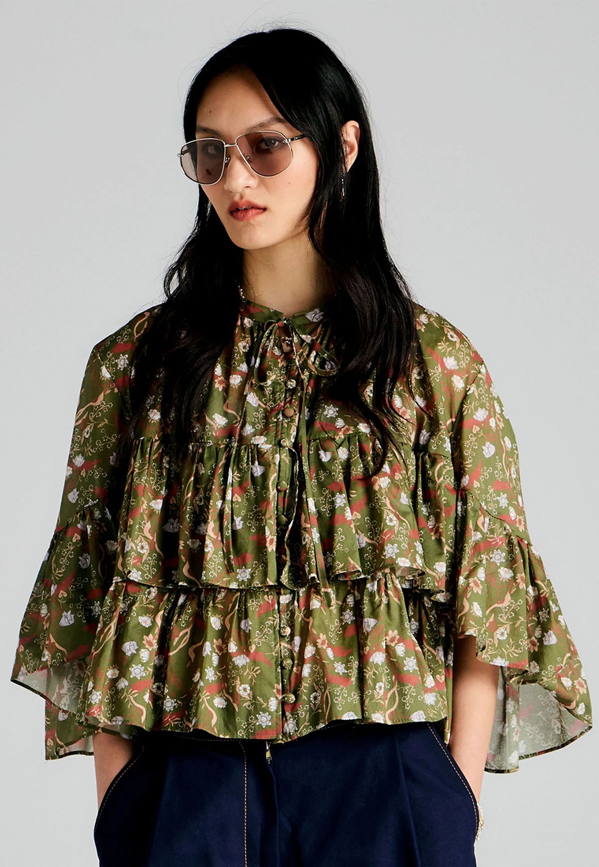 A woman wearing a Karen Walker green tiered floral blouse with a red and white floral pattern, paired with dark blue pants and gold-framed sunglasses, against a solid gray background.