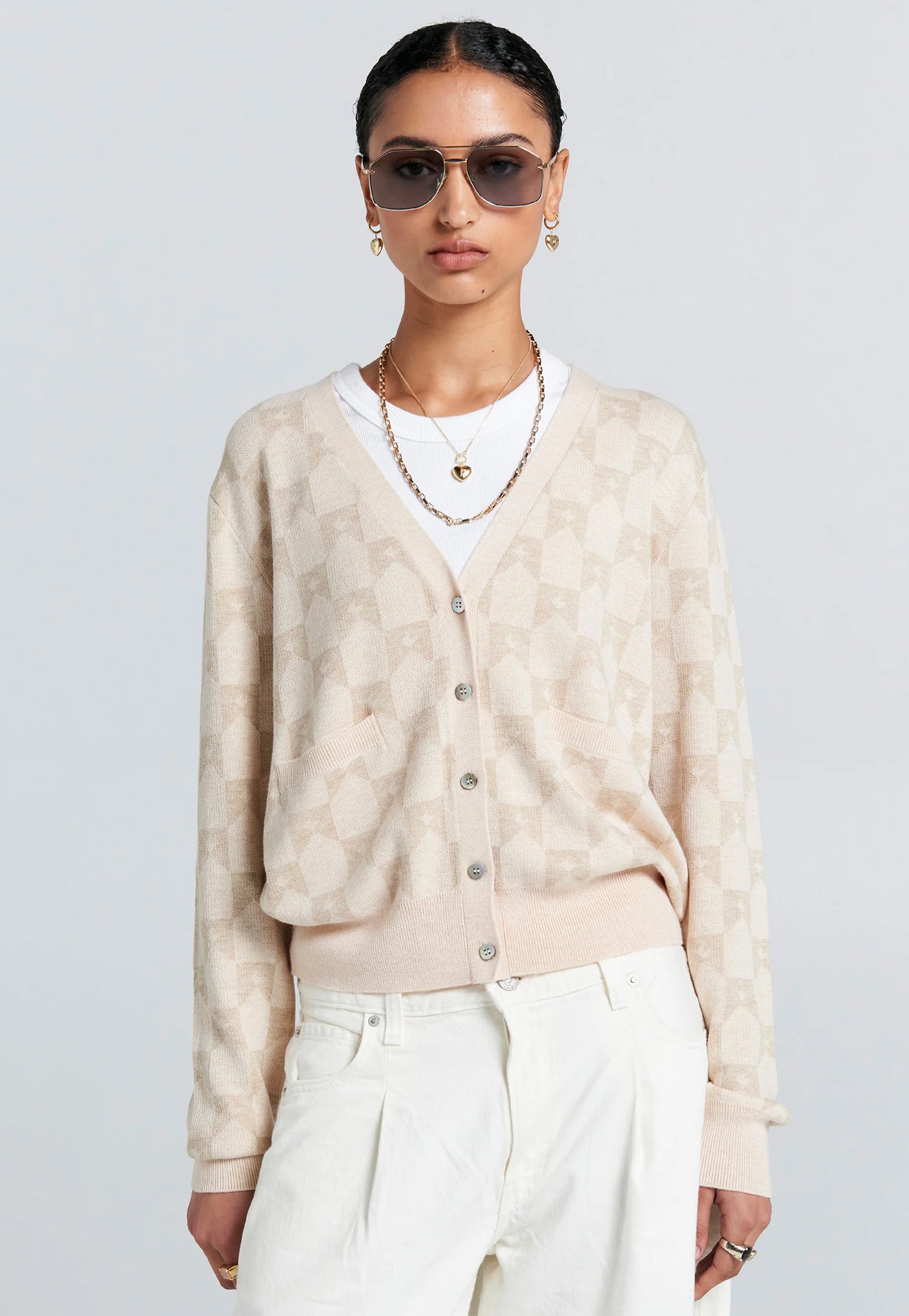 Woman wearing a beige cardigan, white shirt, and white pants against a light gray background