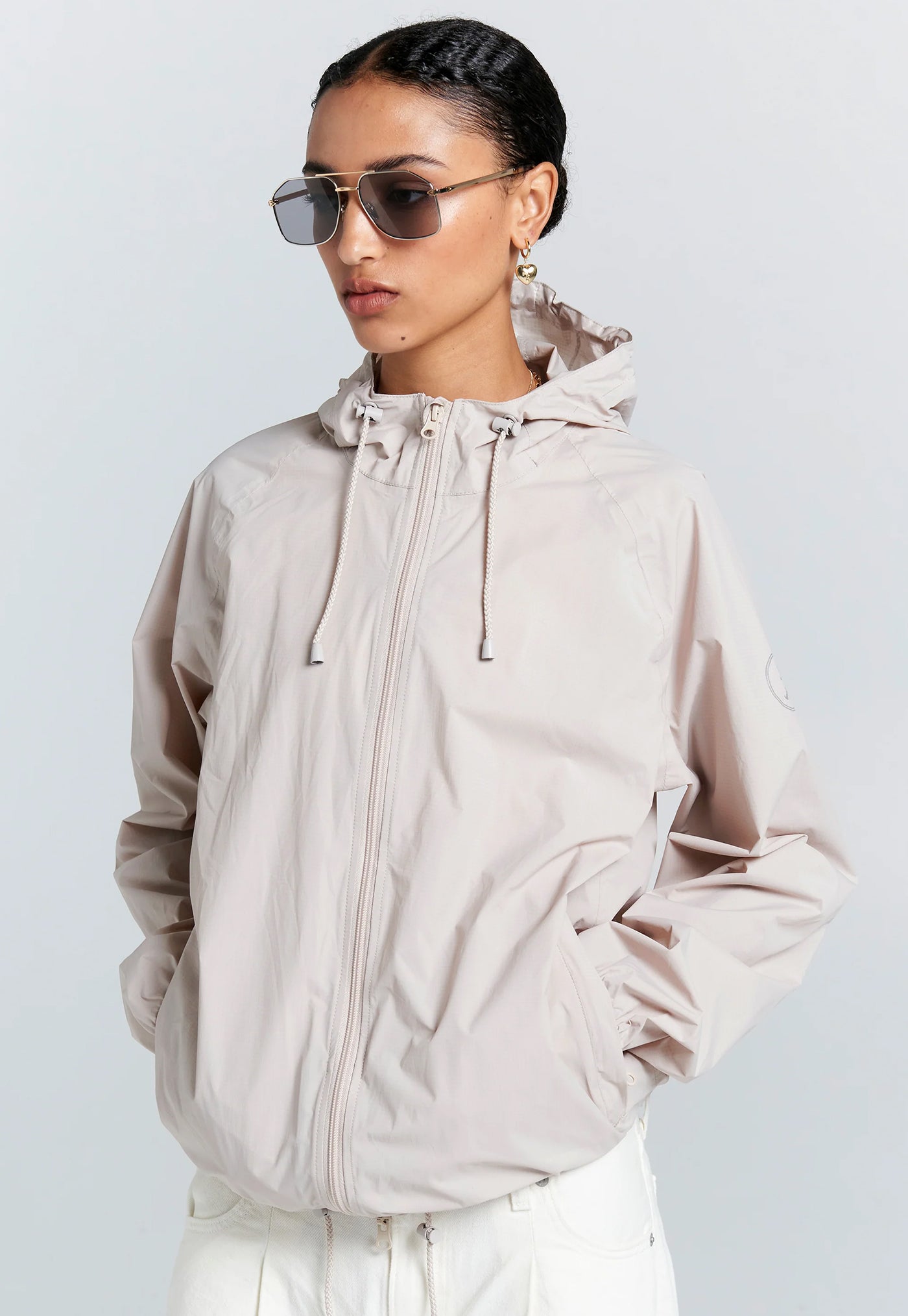 Person wearing a beige hooded jacket and sunglasses on a light gray background