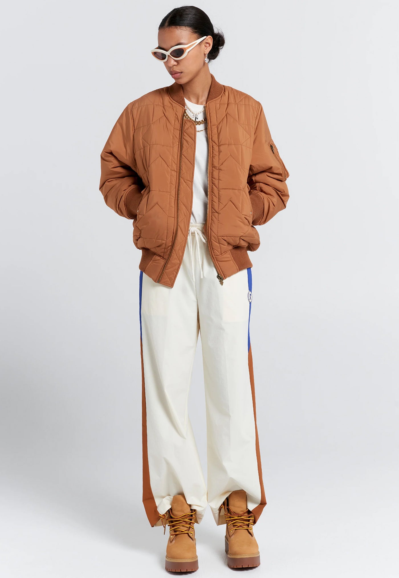Person wearing a brown quilted jacket, white pants with blue stripes, and brown boots on a light gray background