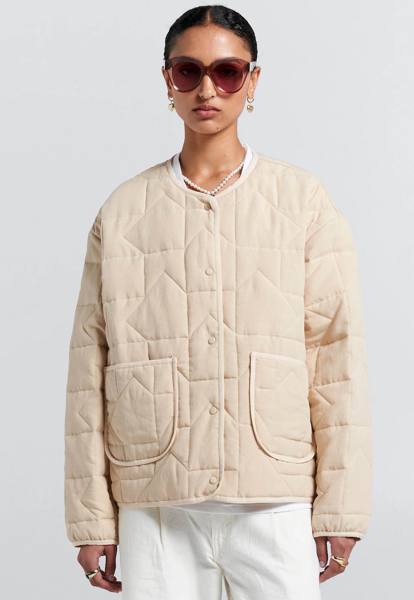 Woman wearing a beige quilted jacket and sunglasses on a light gray background