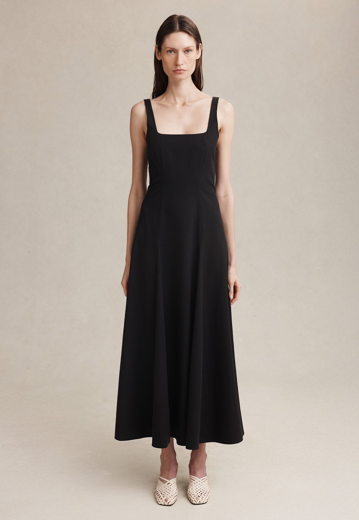 A woman modeling a black sleeveless maxi dress with a square neckline and wide shoulder straps, paired with light beige woven sandals, against a neutral beige studio backdrop.
