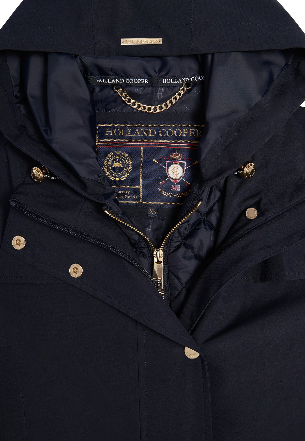 Close-up of a navy blue hooded jacket by Holland Cooper, showing the inner collar with a gold chain and brand label, plus gold-tone snap and zipper details.